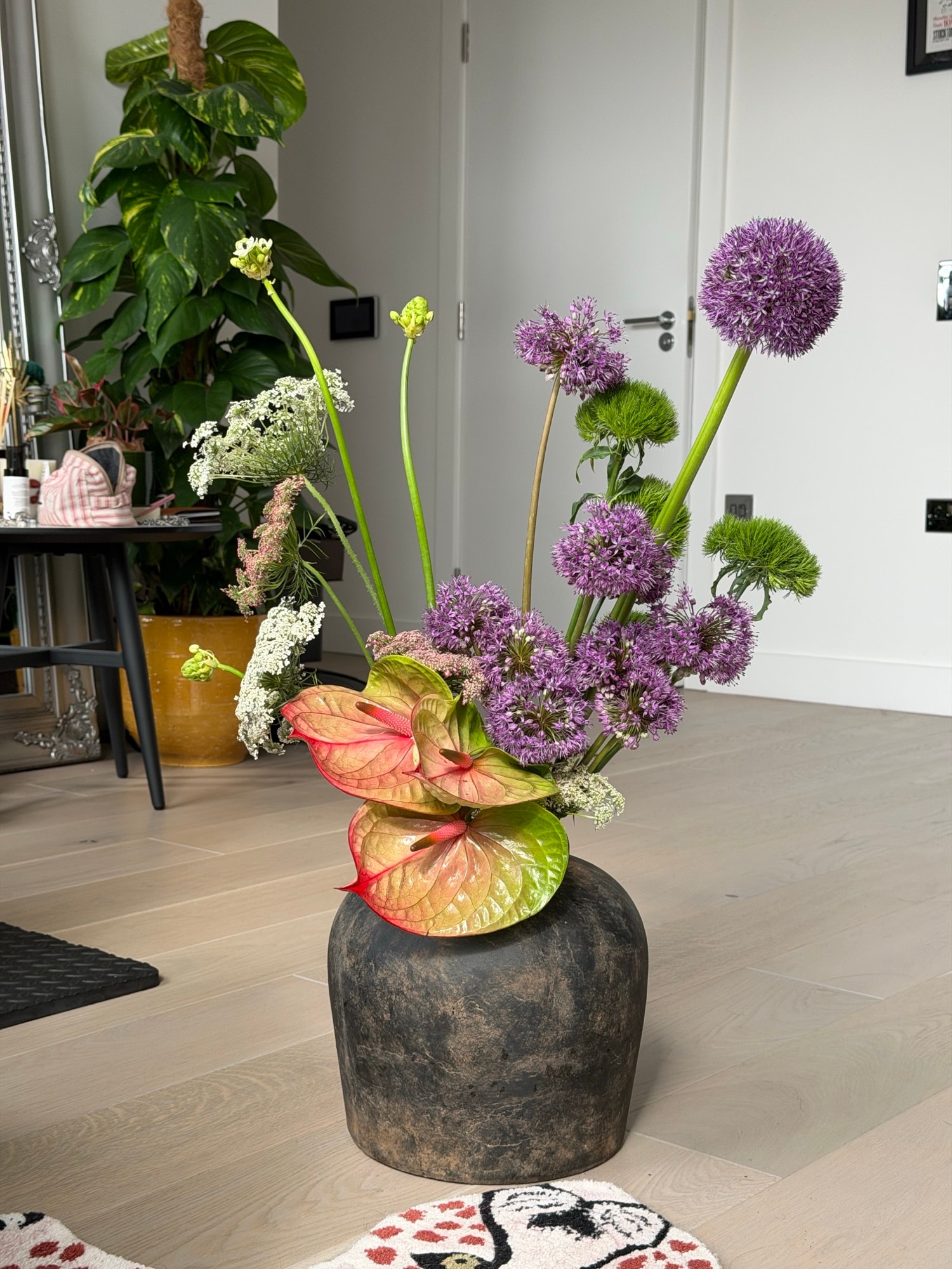Tall sculptural arrangement of purple alliums and variegated anthuriums in a dark ceramic vase on a wood floor