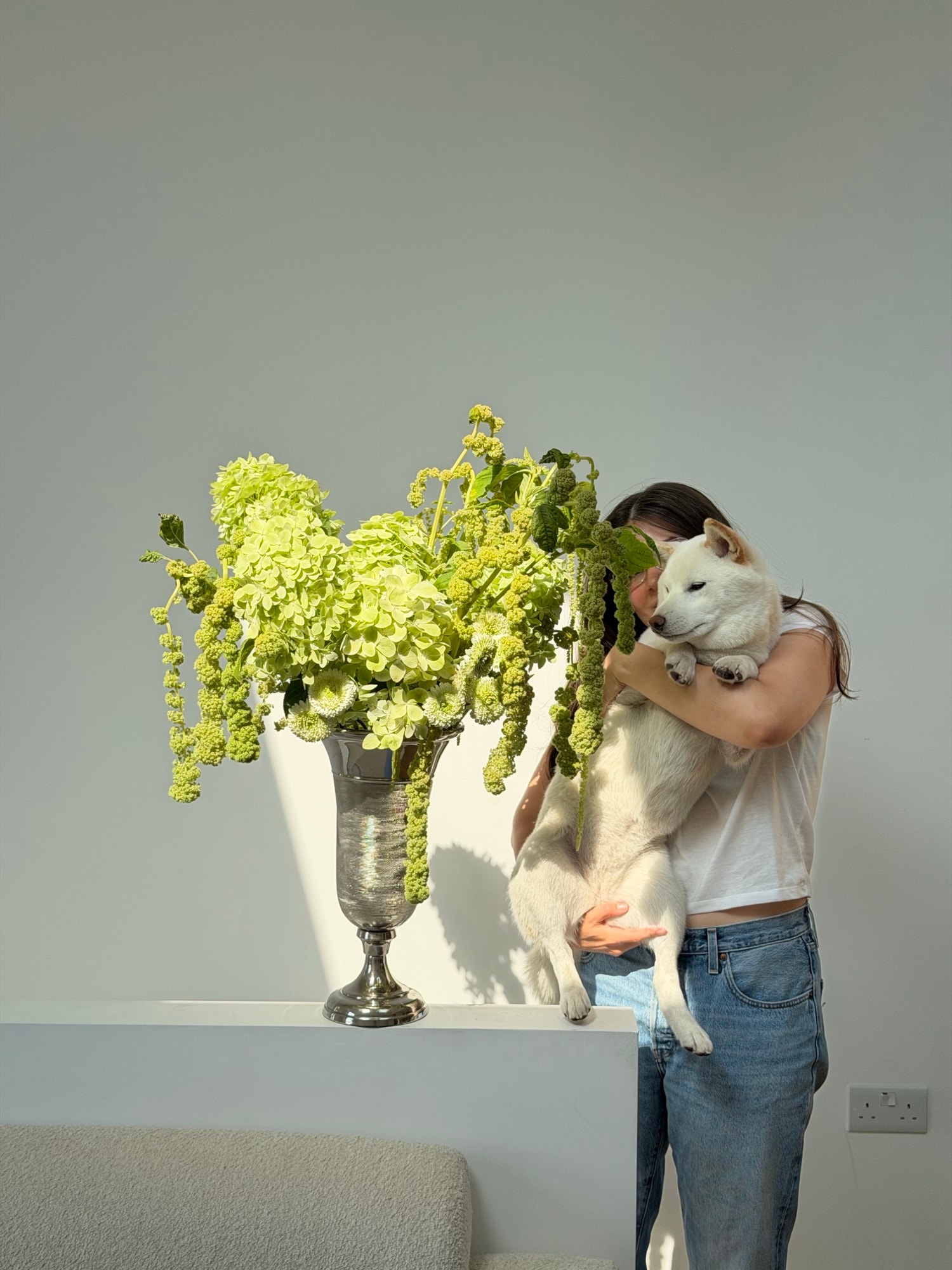 Founder hugging a fluffy white dog behind a sculptural green arrangement in a silver vase