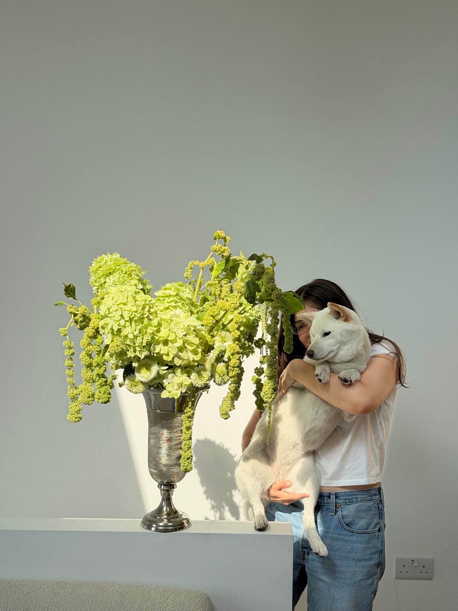 Founder holding Misha while looking toward overflowing green flowers in a silver vase