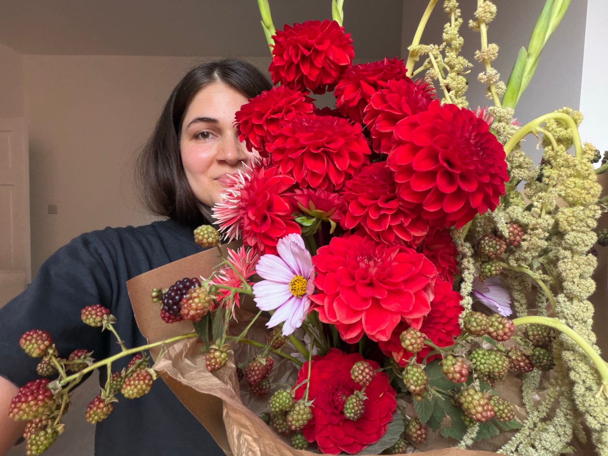 Woman peeking over a kraft-wrapped bouquet of red dahlias, pink cosmos, and blackberry stems