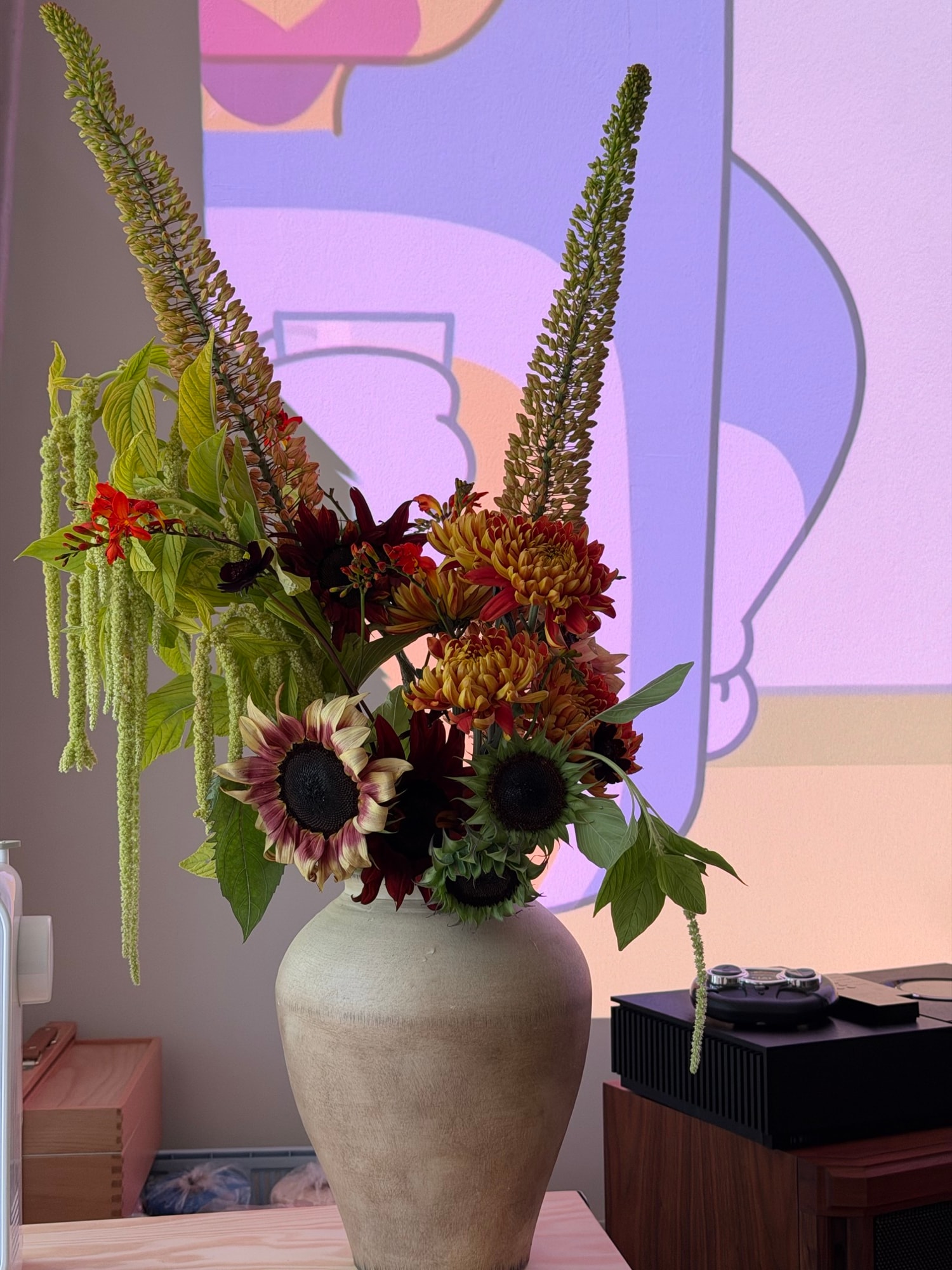 Tall off-white vase with sunflowers, bronze chrysanthemums, and fuzzy green spikes beside a projected landscape